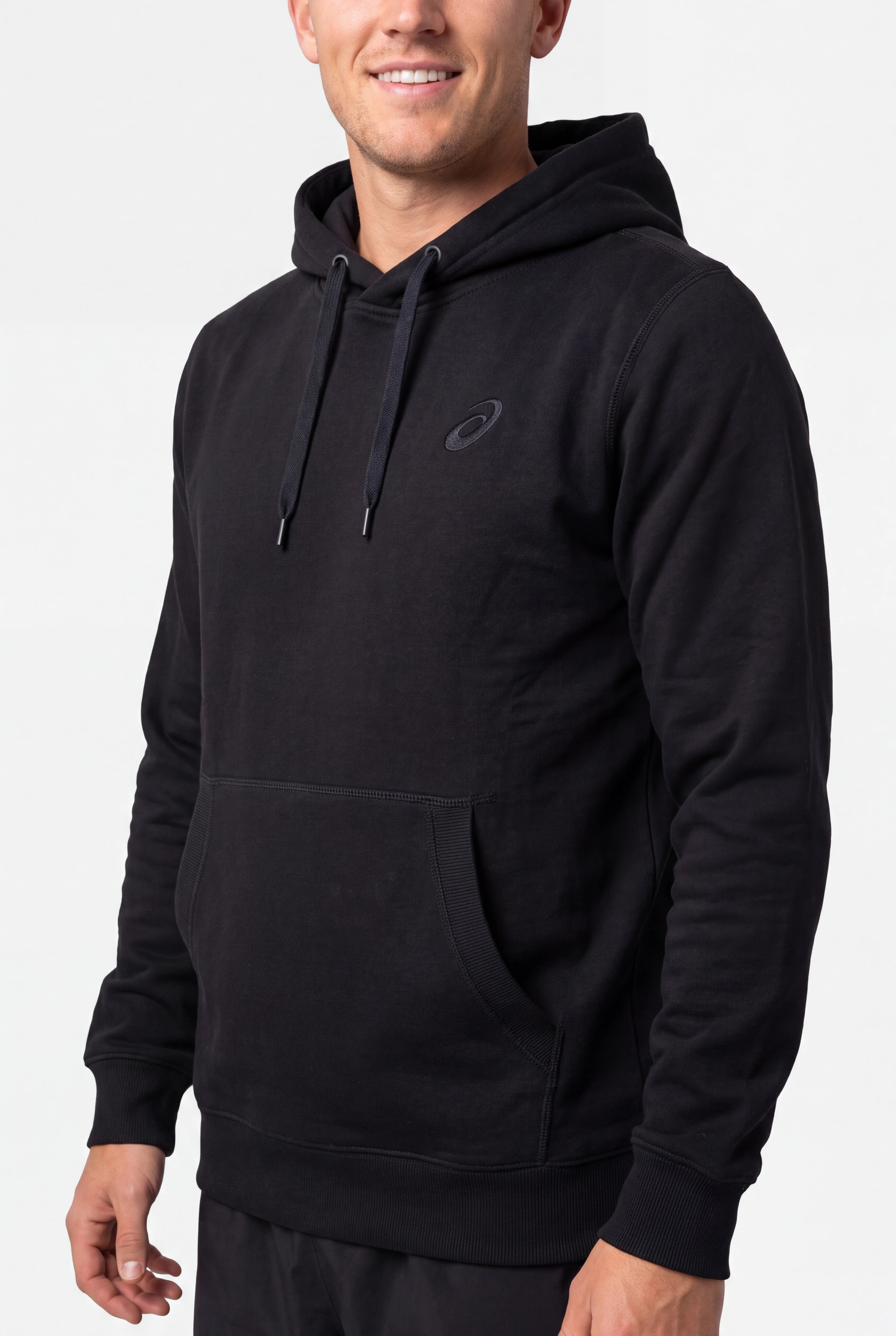 A man wearing a black pullover hoodie with a kangaroo pocket and a small tonal Asics logo on the left chest, photographed against a light gray studio background.