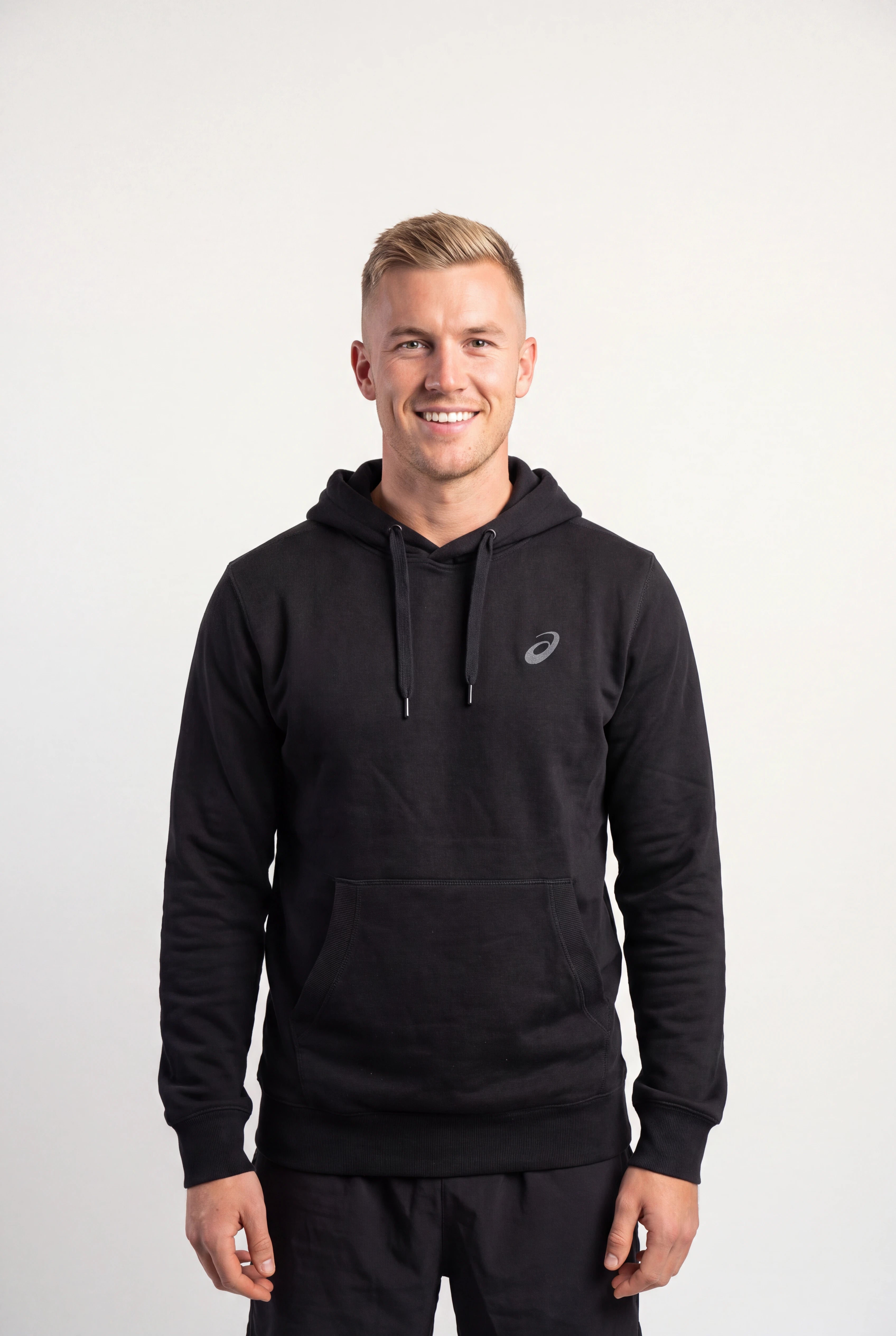 A smiling man wearing a black Asics pullover hoodie with a small white logo on the left chest, standing against a plain light gray studio background.