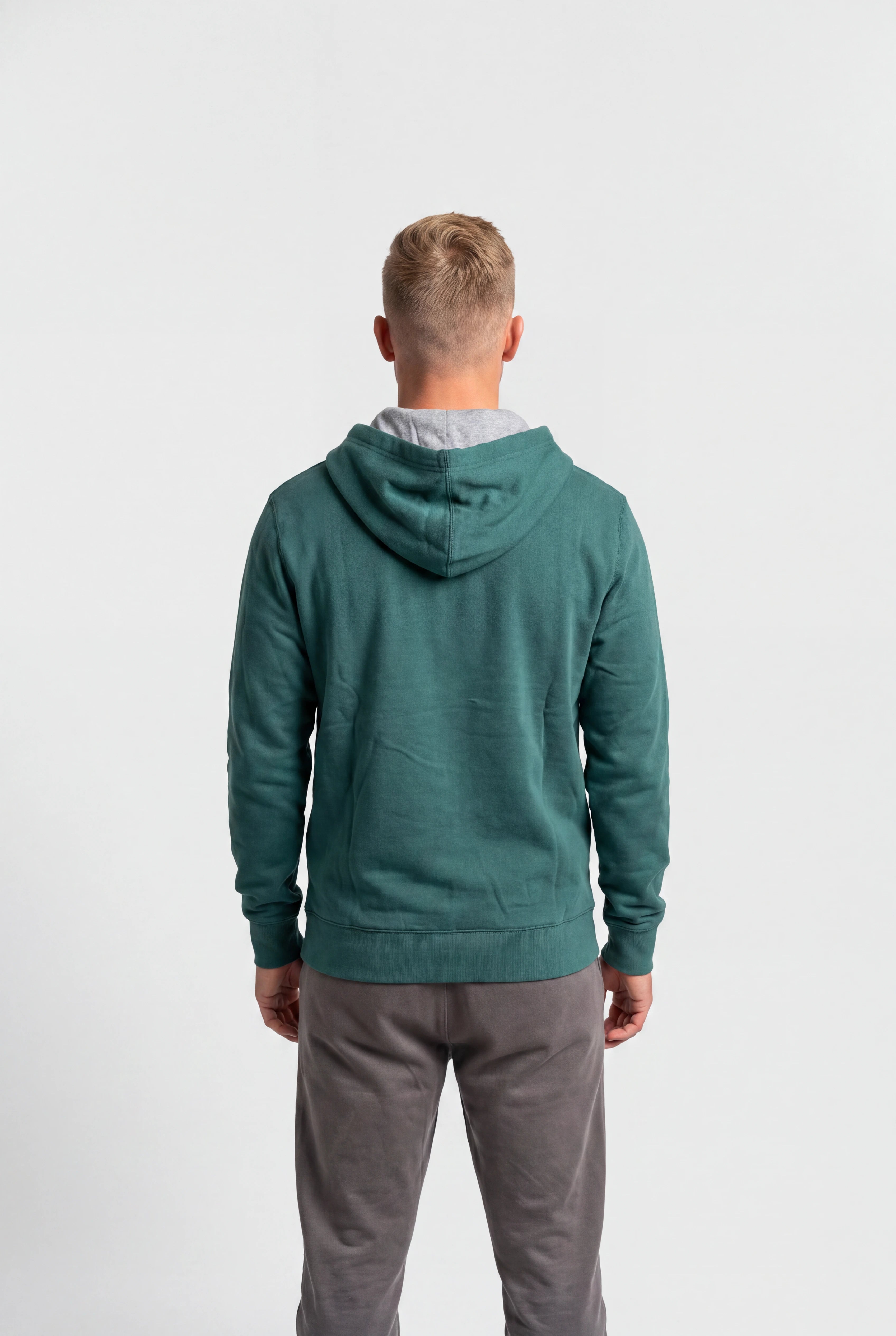 A man viewed from behind wearing a sage green hooded sweatshirt and gray sweatpants against a plain white studio background.