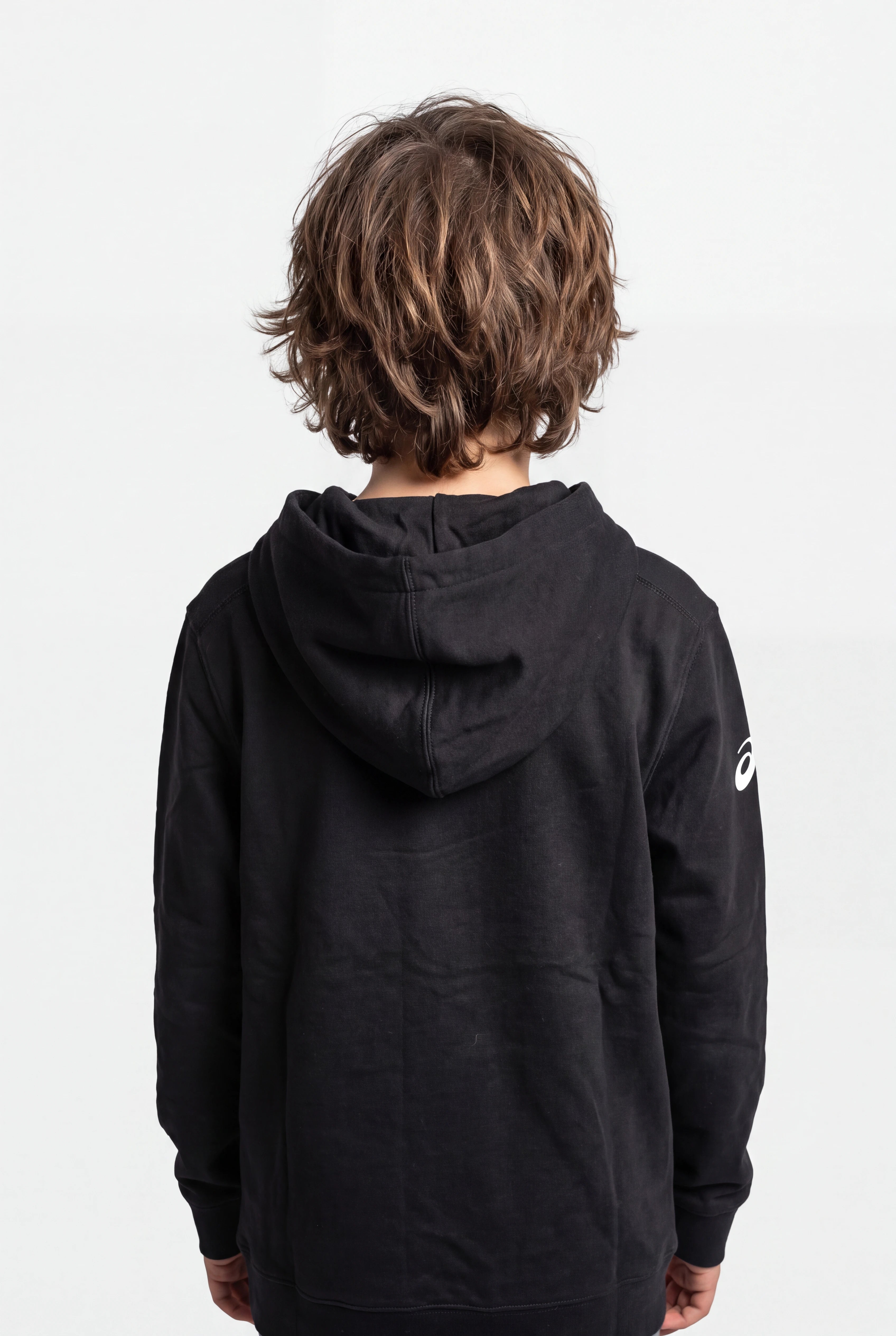 A child viewed from behind wearing a black hooded sweatshirt with a small white Asics logo on the left sleeve, set against a plain light gray studio background.