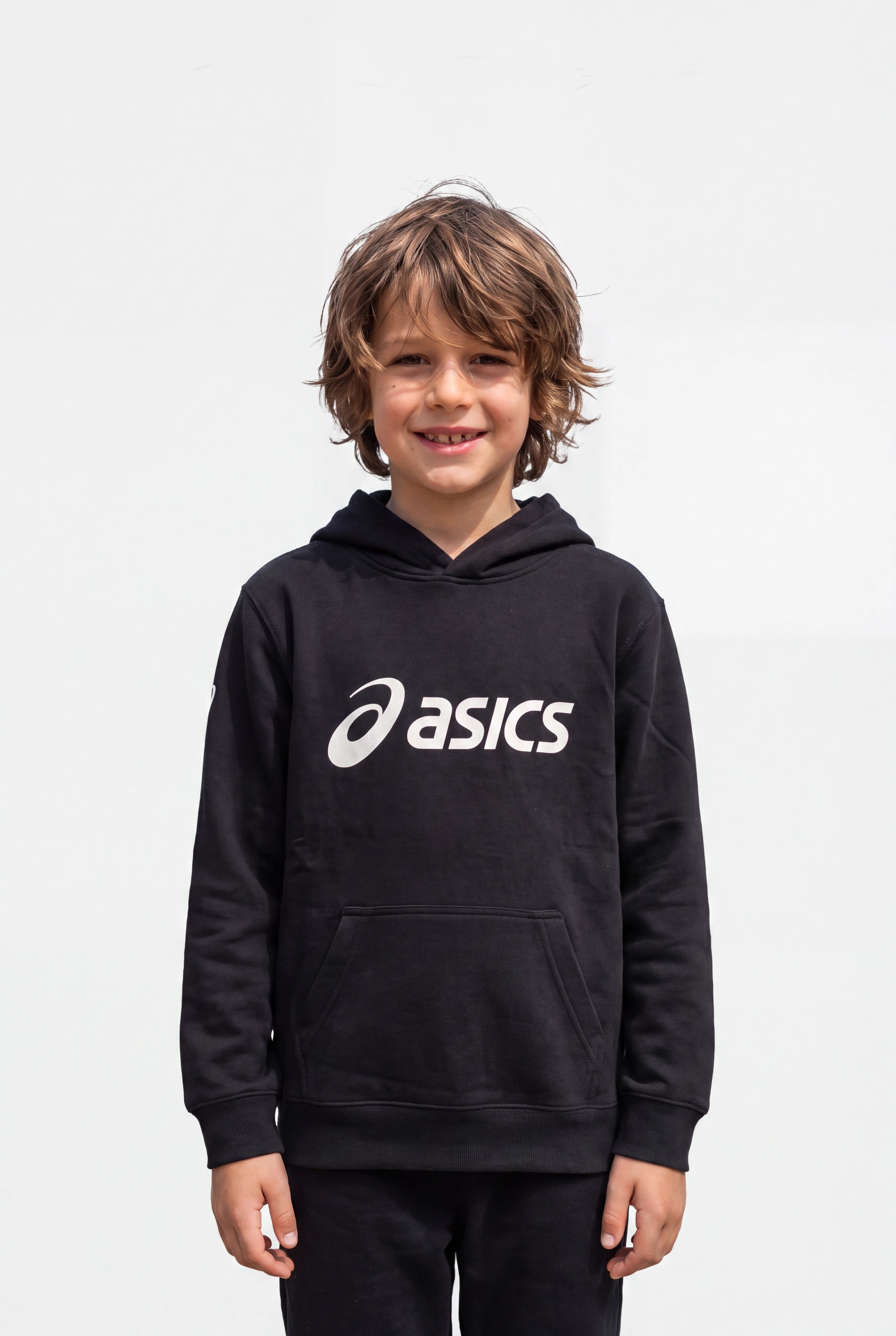 A young boy with wavy brown hair smiling, wearing a black Asics pullover hoodie with a white logo on the chest, set against a plain light gray background.
