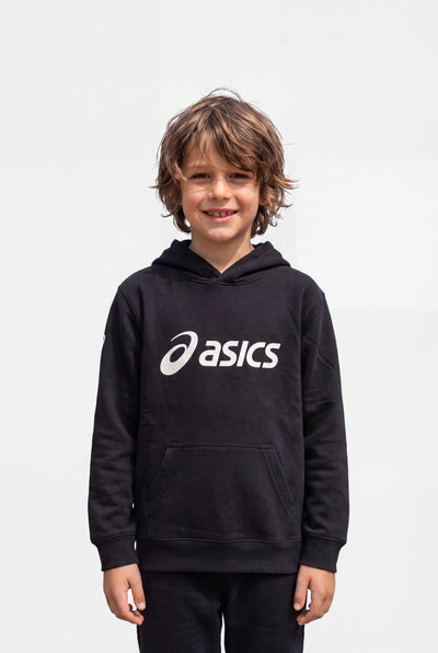 A young boy with wavy brown hair smiling, wearing a black Asics pullover hoodie with a white logo on the chest, set against a plain light gray background.