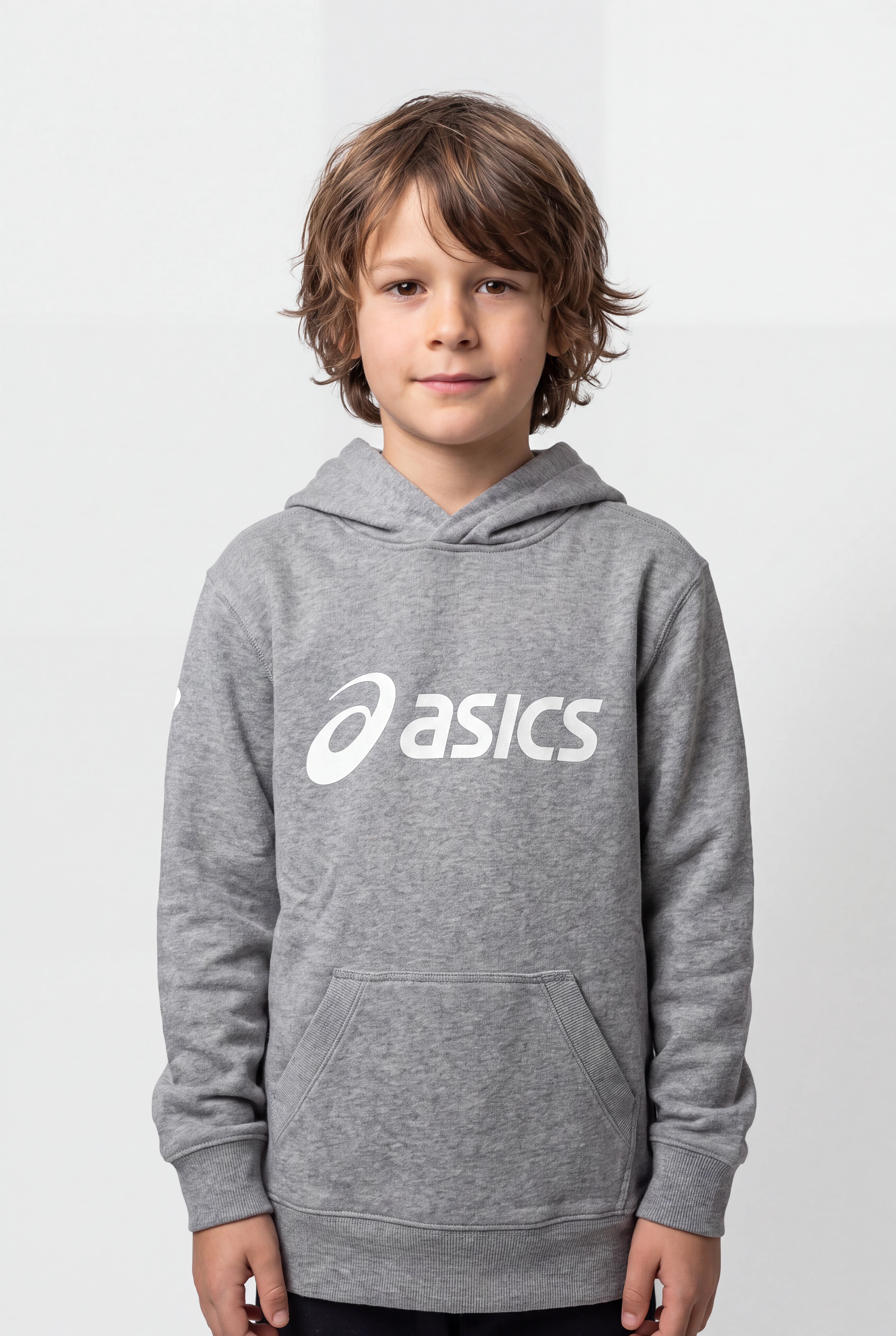 A young boy with wavy brown hair wearing a heather gray Asics pullover hoodie with a white printed logo on the chest, standing against a light gray studio background.