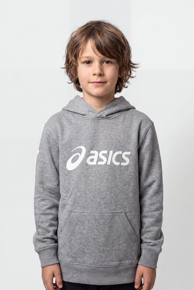 A young boy with wavy brown hair wearing a heather gray Asics pullover hoodie with a white printed logo on the chest, standing against a light gray studio background.