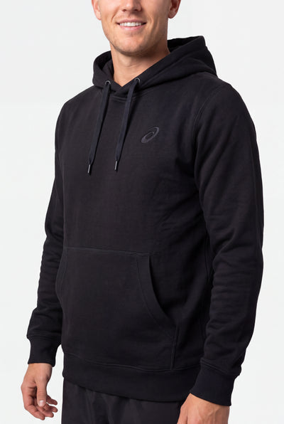 A man wearing a black pullover hoodie with a kangaroo pocket and a small tonal Asics logo on the left chest, photographed against a light gray studio background.