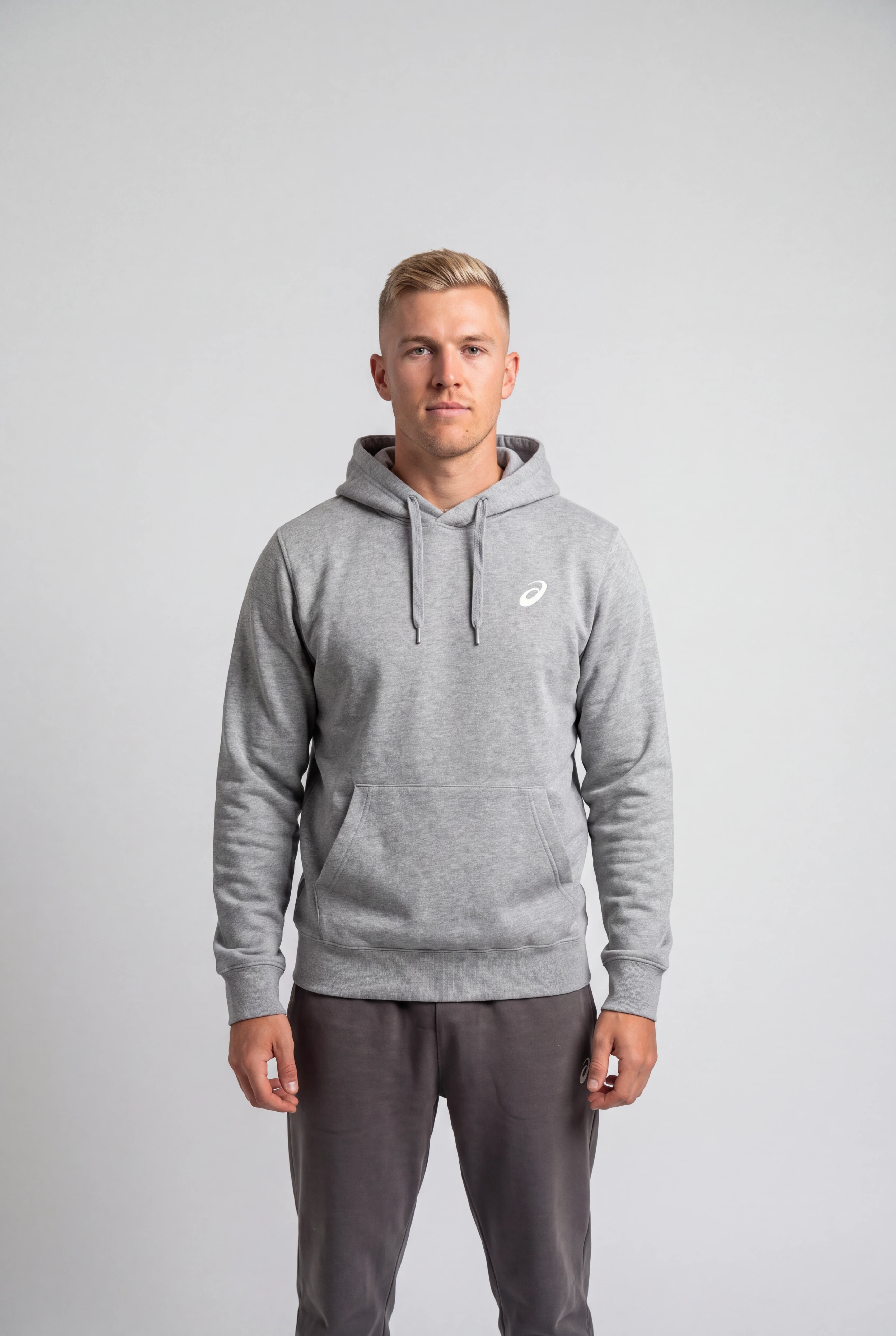 A man wearing a light gray heather pullover hoodie with a small white Asics logo on the left chest, paired with dark gray sweatpants, standing against a light gray studio background.