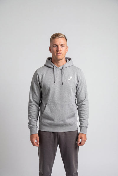 A man wearing a light gray heather pullover hoodie with a small white Asics logo on the left chest, paired with dark gray sweatpants, standing against a light gray studio background.