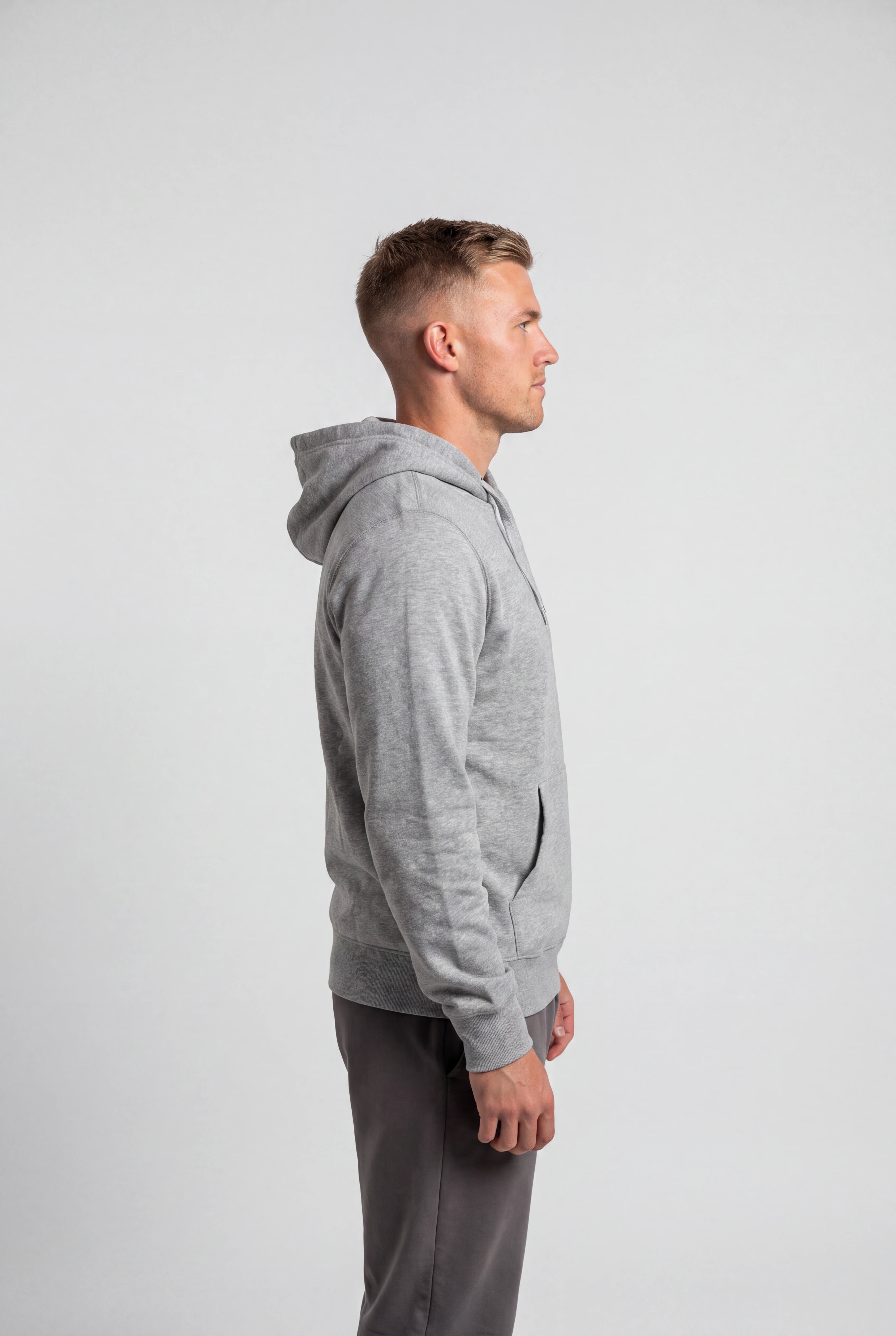 A man in profile wearing a light gray heathered zip-up hoodie and dark gray sweatpants, standing against a solid light gray studio background.