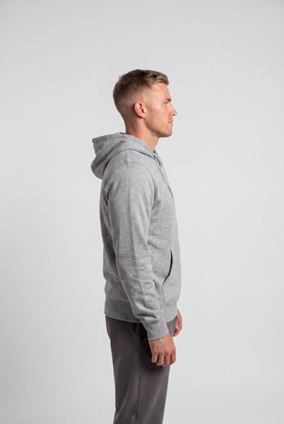 A man in profile wearing a light gray heathered zip-up hoodie and dark gray sweatpants, standing against a solid light gray studio background.