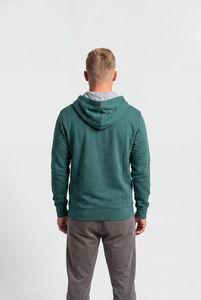 A man viewed from behind wearing a sage green hooded sweatshirt and gray sweatpants against a plain white studio background.