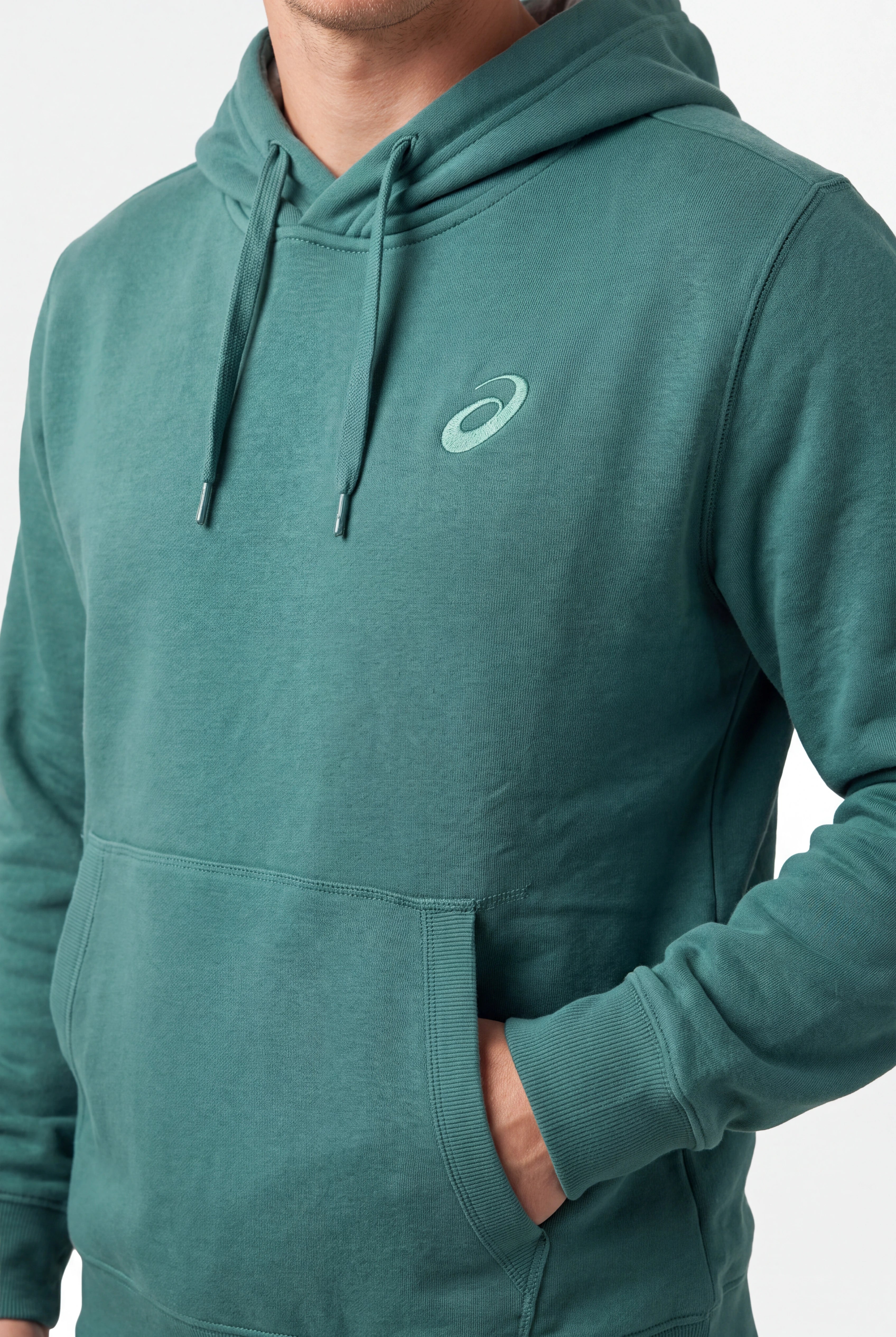 A close-up of a person wearing a teal-green pullover hoodie with a small white Asics logo on the left chest, set against a plain light gray background.