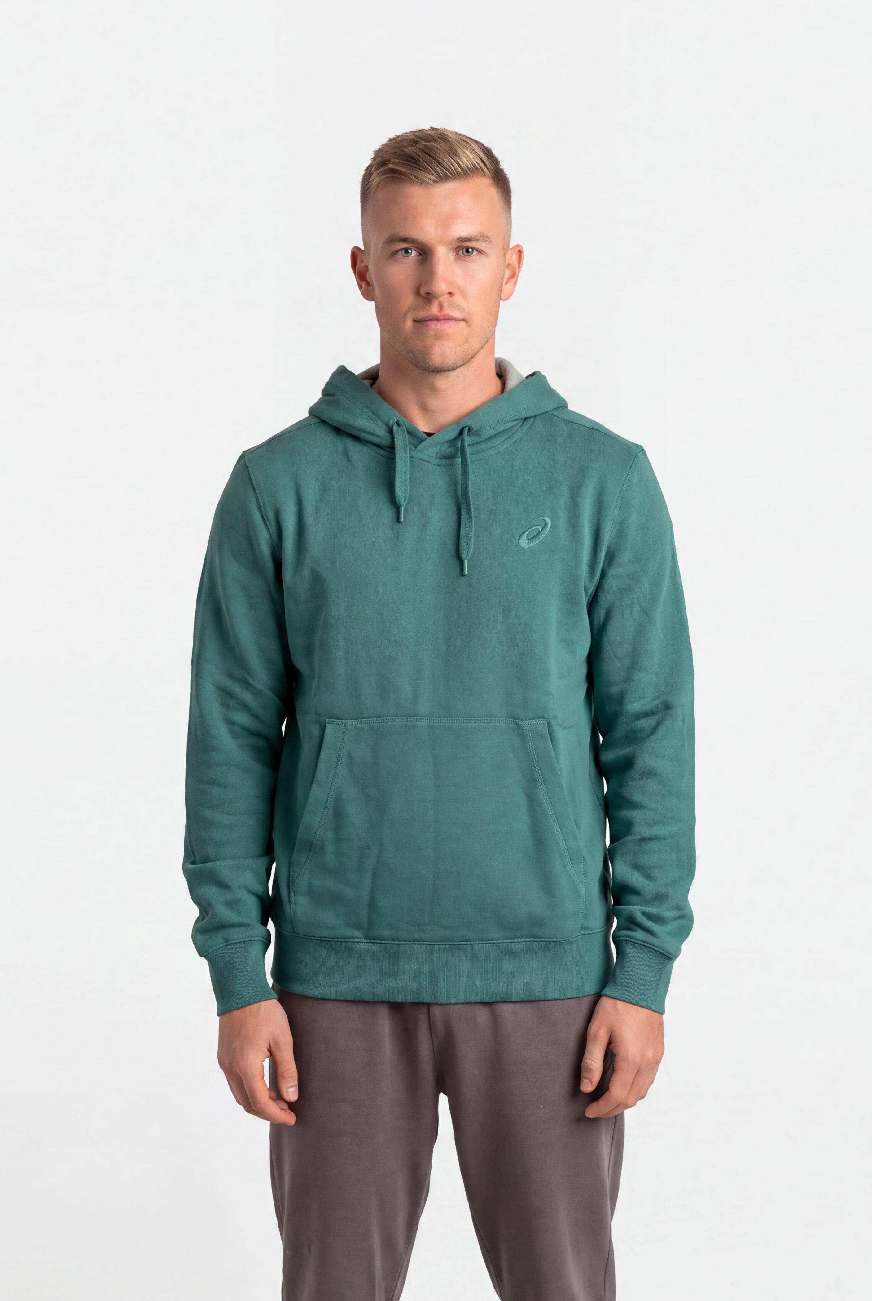 A man wearing a teal green pullover hoodie with a small white embroidered logo on the left chest, paired with gray sweatpants, standing against a plain white studio background.