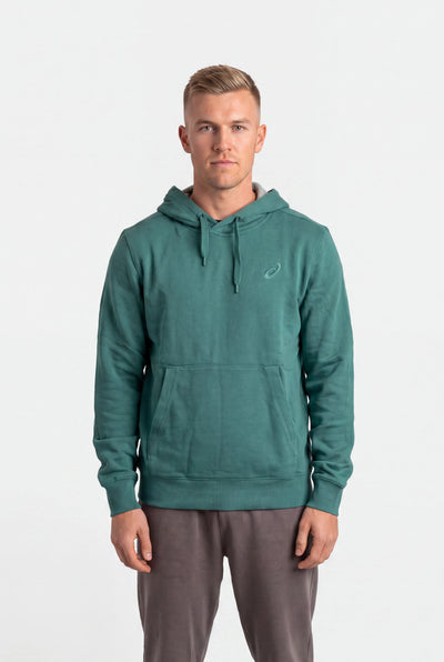 A man wearing a teal green pullover hoodie with a small white embroidered logo on the left chest, paired with gray sweatpants, standing against a plain white studio background.