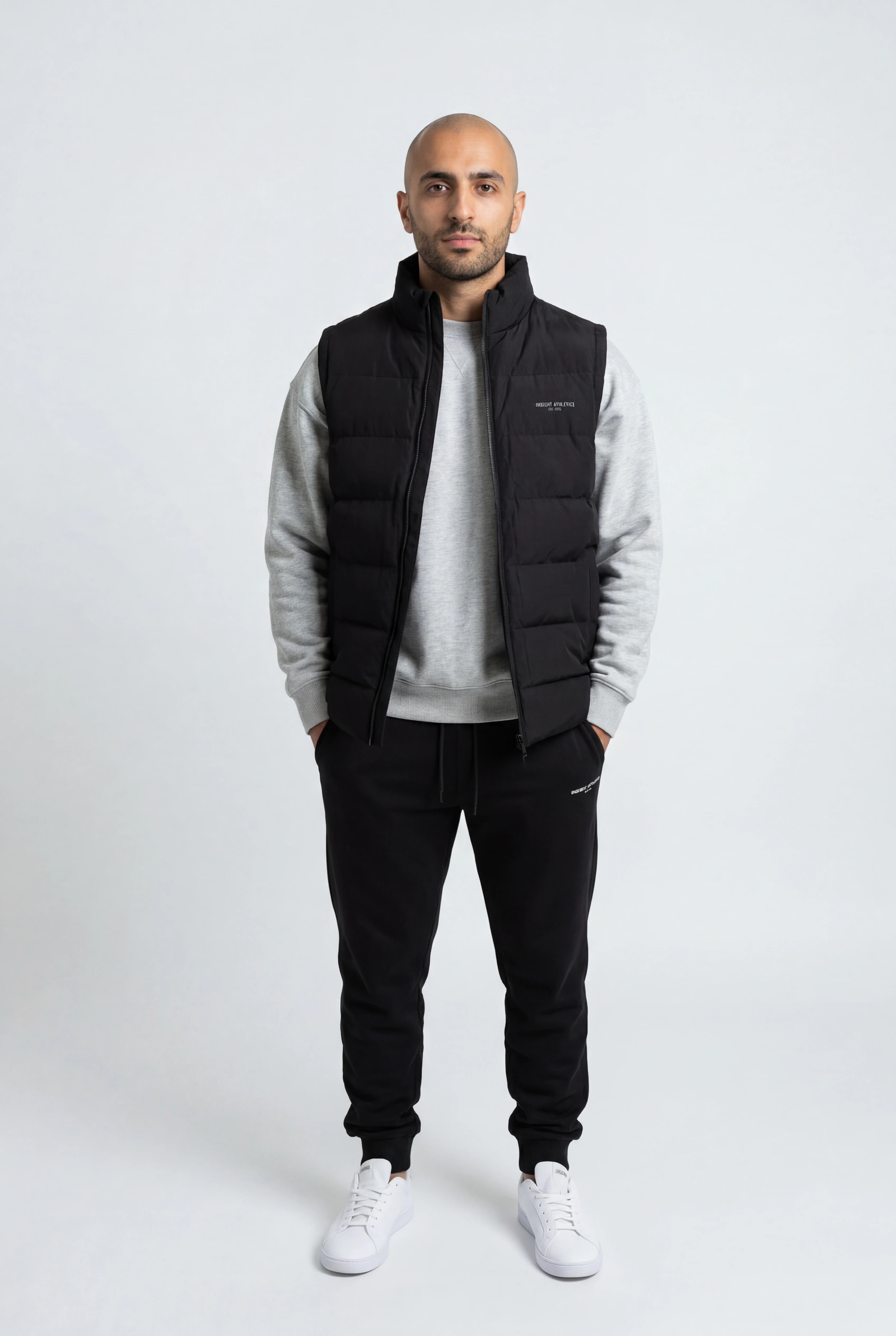 Man wearing a black puffer vest over a gray sweater with black pants on a white background