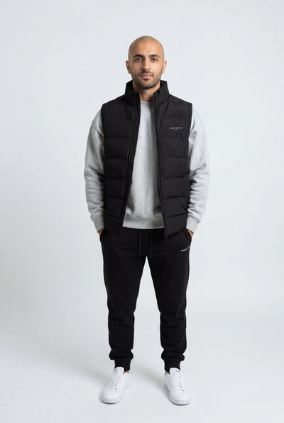 Man wearing a black puffer vest over a gray sweater with black pants on a white background